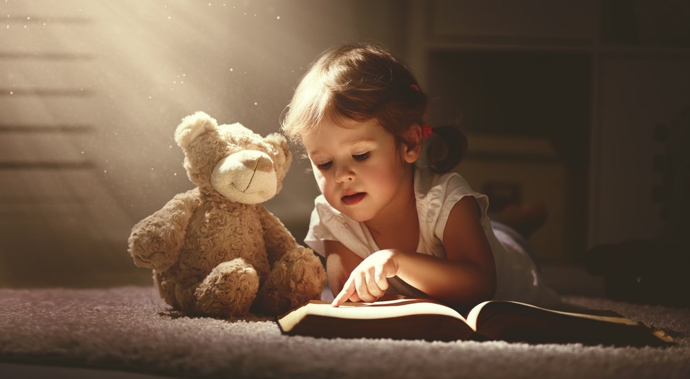 A young child reading a book next to a teddy bear