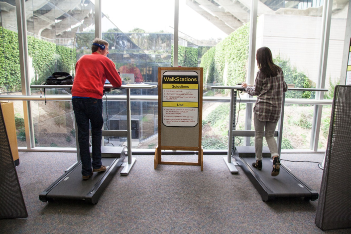 Two students walking on treadmills side by side while facing the window.