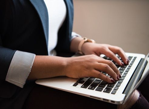 Image of a Black person from the chest down to their knees. They are wearing a navy blue blazer with rolled up sleeves, a rose gold watch on their left wrist, and their nails are painted a burgundy color. Their hands are typing away at a Mac laptop which is positioned over their thighs. They are wearing ripped jeans and there is a tear on their right knee, peaking through at the bottom right corner of the image. The background behind them is a mauve color.