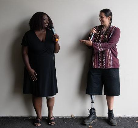 An Indigenous Two-Spirit person and a Black woman face each other and gesture while smiling and speaking into microphones. They are both wearing prosthetic legs.