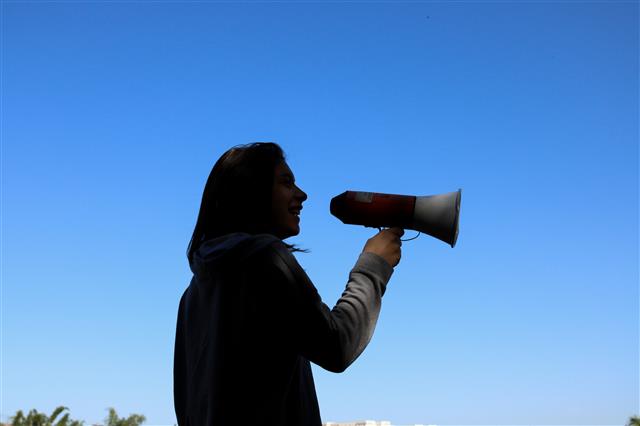 A photo of a person with long hair and a grin holding a megaphone up to their mouth. They are wearing a grey long sleeve shirt. The background is the blue sky with some green leaves peeking through at the bottom left corner of the photo.