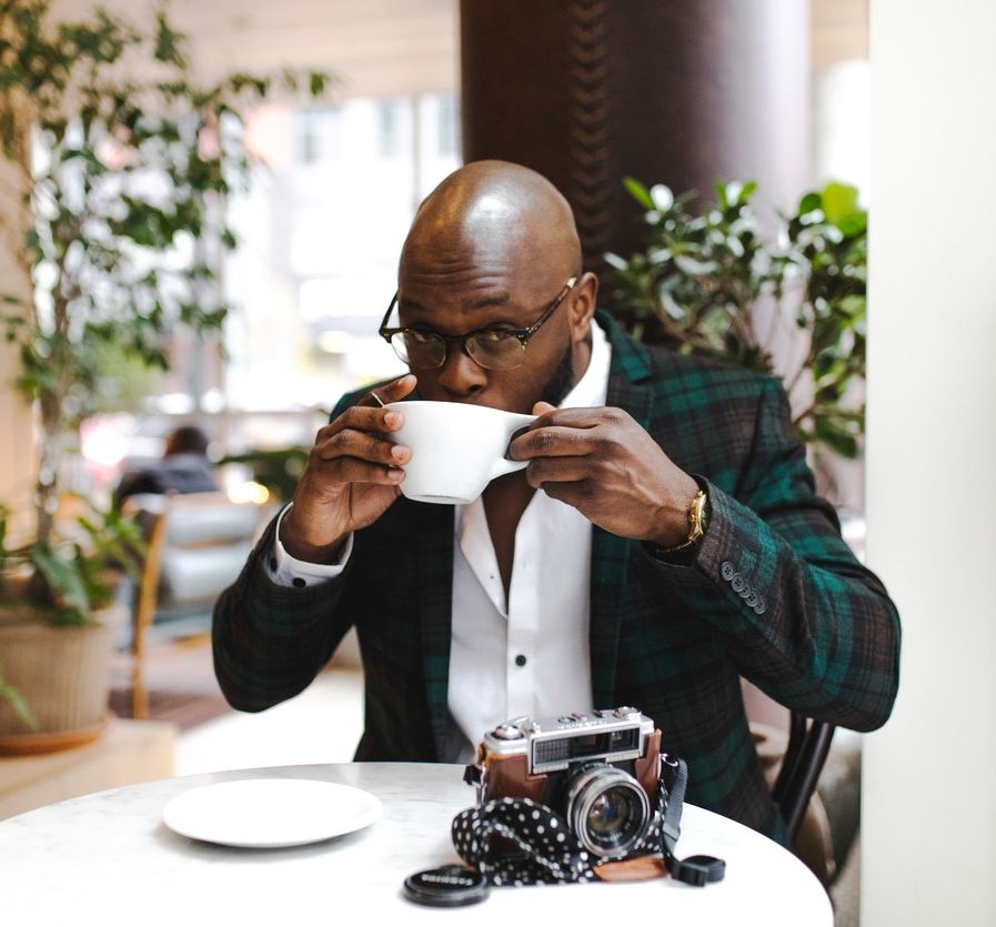 A dark-brown Black man with a button-up white shirt and a tweed greed and black coat on is looking up through his glasses, subtly smiling, while sipping from a cup. In the background is a wide brown pillar and plants on either side. There is a table in front of the man and on top of it is the cup's saucer and a vintage-looking camera. 