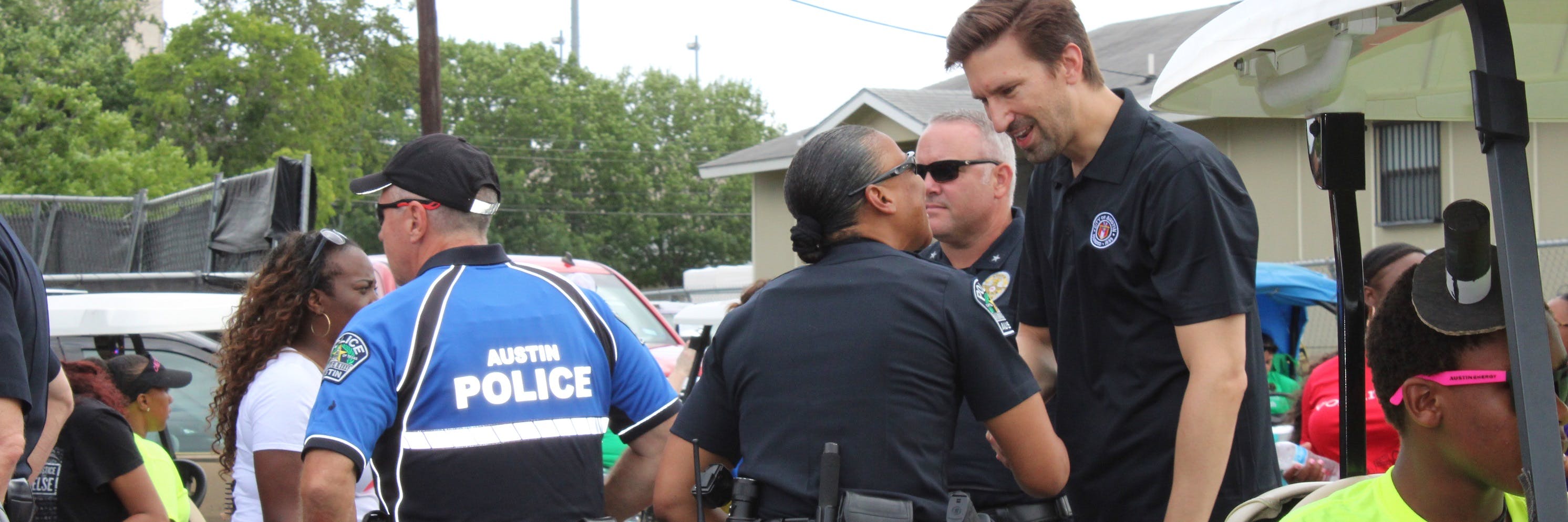A photo of City Manager Spencer Cronk shaking a police officer's hand and speaking to them. There are a few police officers photographed along with some civilians. It is day-time and people are outdoors.