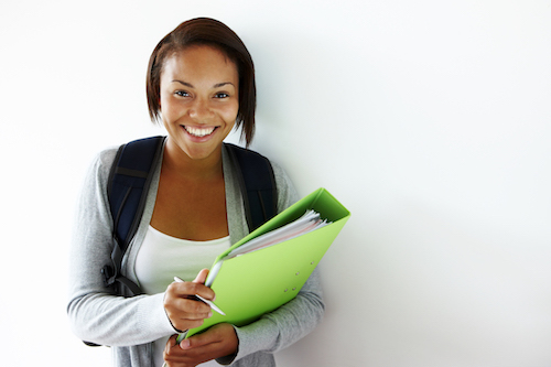 A person who looks like a student, wearing a backpack and holding a big green binder with a pen in hand, is smiling at the camera. There is natural light on the person. The background is a white wall.