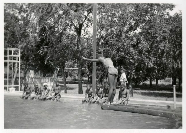 Black and white photo from 1938: A group of black children watching a youth prepare to dive from the diving board into the public pool at Rosewood Park.