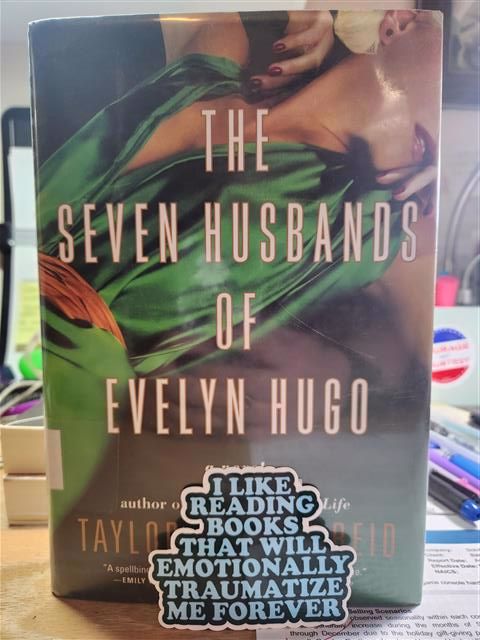 A photo of "The Seven Husbands of Evelyn Hugo" by Taylor Jenkins Reid, which has a white woman with blond hair, deep red nails, and a stunning green dress provocatively lying on her back. Her face above her red lips is hidden.  In front of the book, I've placed a blue sticker that reads "I like reading books that will emotionally traumatize me forever."