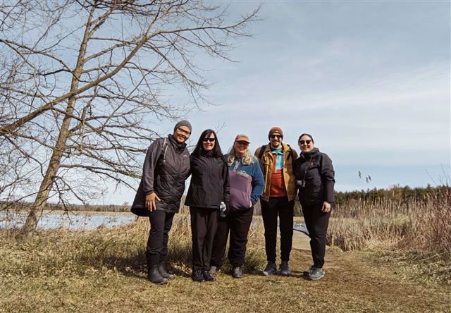 A picture of 5 adults wearing outdoor gear standing in front of a grey sky. They are all facing the camera and smiling. In the background there is a bit of lake, and some trees. 