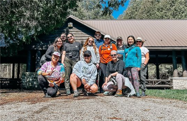 A picture of 5 adults wearing outdoor gear standing in front of a grey sky. They are all facing the camera and smiling. In the background there is a bit of lake, and some trees. 