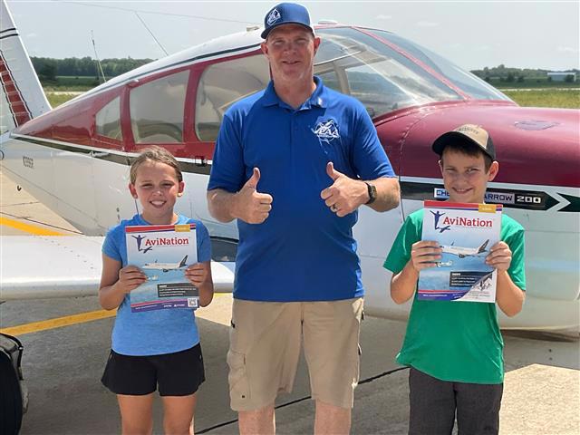 2 children standing on either side of adult male in front of a GA plane