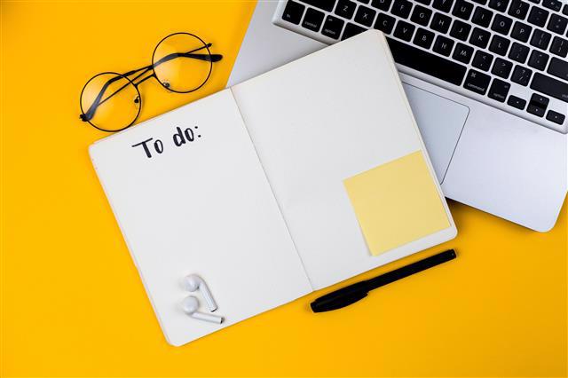 [Image Description: Yellow background with open laptop, glasses, and an open notebook in the foreground. Airpods and a blank sticky note are sitting on the notebook; top of the first page reads: To do."