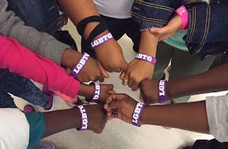 Hands meet in the middle each wearing LGBTQ purple bracelets.