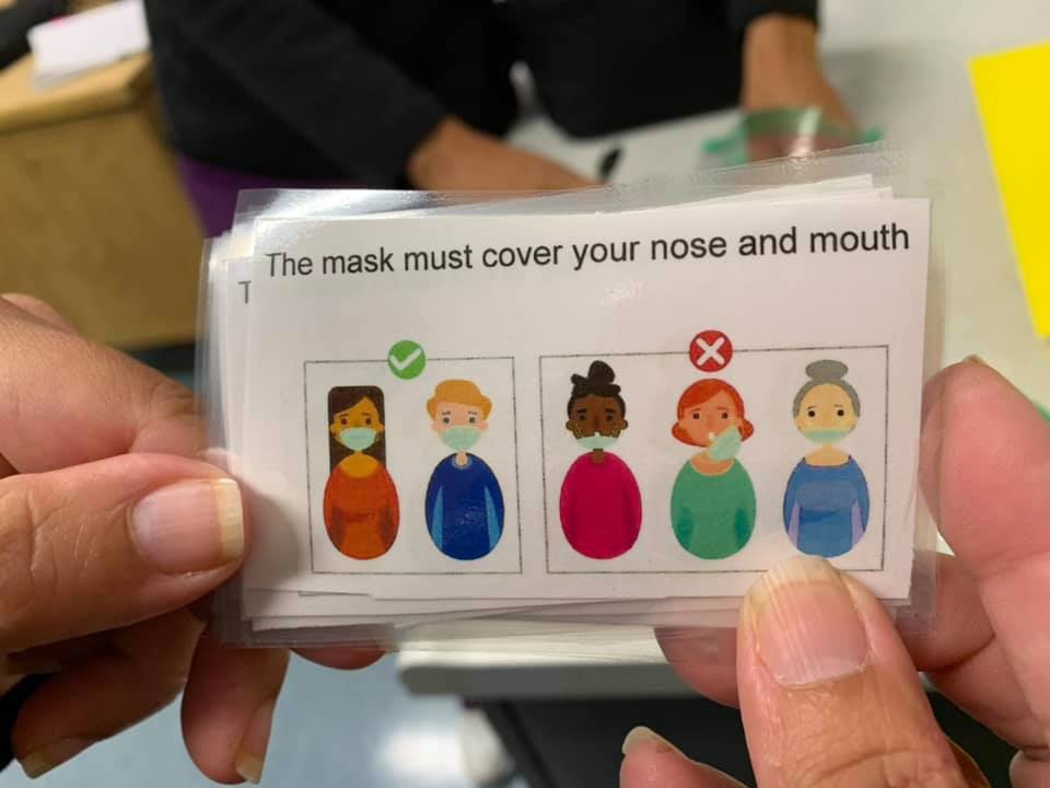 Two hands hold a card that reads "The mask must cover your nose and mouth"