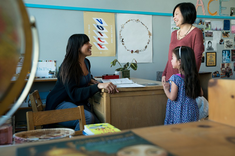 A female teacher speaks with a mom and young female student in a classroom.