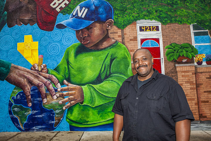 A black man stands in front of a colorful mural featuring a child with a globe