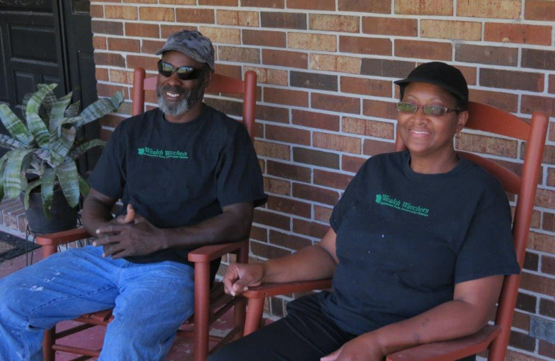 A black man and a black woman sit in wooden chairs in front of a brick building.