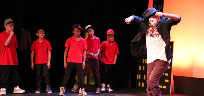Five boys wearing red shirts watch another boy wearing a half mask dancing on stage