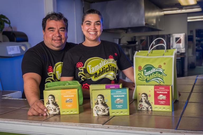 Two Latinx individuals stand in their start-up kitchen with their boxes of tamales