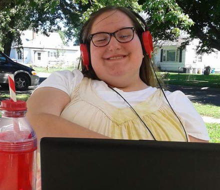 A girl wearing a yellow dress has red headphones and stares at her laptop outside