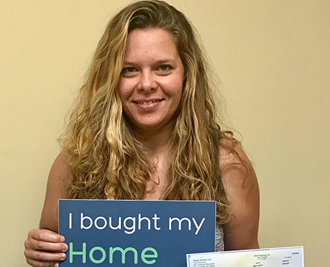 A woman with blonde hair holds up a check and a sign that says: I bought my home with Origin SC