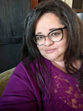 Tigress Osborn is a middle-aged fat Black mixed-race woman with long, dark hair and freckles. She is wearing black framed glasses and a purple top.