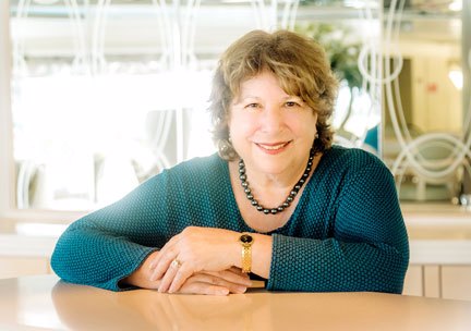 A photo of Barbara Altman Bruno PhD; a smiling fat Jewish woman with short hair, wearing a green dress, a bead necklace and a gold watch, sitting at a table with mirrors in the background