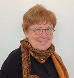 A photo of Esther Rothblum PhD, a smiling fat white womxn with short red hair, wearing round-framed glasses, a black sweater with a print scarf around her neck