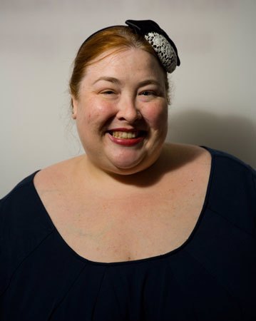 Portrait pic of Cat Pausé, PhD, a smiling fat white womxn with red hair pulled back and wearing a black dress and a black and white fascinator.