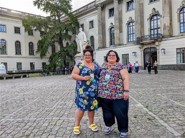 NAAFA Board Members Tigress Osborn and Elaine Lee are posing in front of historic Senate Hall at Humboldt University in Berlin. Tigress is a fat Black woman wearing a blue dress with a yellow and white flower pattern. Elaine is a Chinese-American woman wearing black pants and a multicolored top. Both are wearing glasses and smiling.