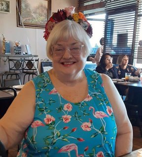 2022 photo of Peggy Howell at her 75th birthday celebration dinner. She is wearing a flamingo print dress, glasses and a floral headband.