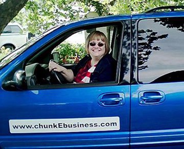 2005 photo of Peggy Howell in the driver's seat of a blue van with a sign reading "www.chunkEbusiness.com" on the side of it. She is smiling and wearing a blue jacket, red, white and blue t-shirt and sunglasses.