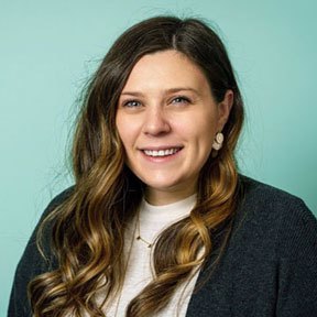 A white female with long brown hair wearing a white shirt and dark gray cardigan. She is smiling in front of a light blue background. (Provided by Samantha Turner)