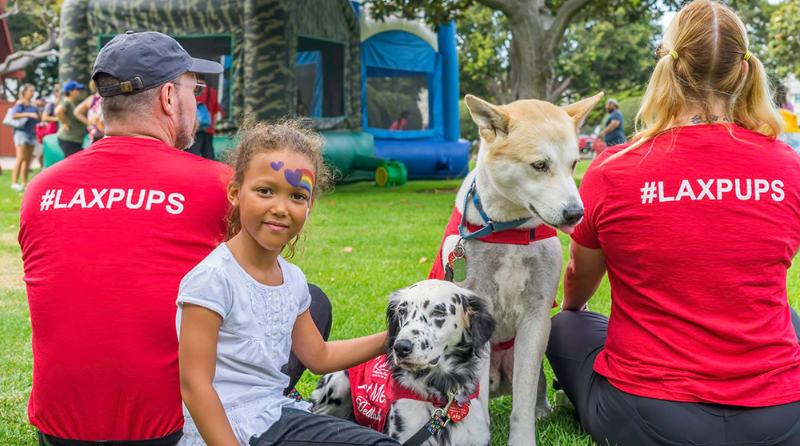 In the center of this photo is a young girl with two dogs from the PUP Program.She has a rainbow with purple hearts painted of the left side of her face and has her hand resting on one of the pups. On either side of her is an adult, and man to her right and a woman to the left, between her and the dogs. They are all sitting on grass and two moon bounces can be seen a few yards away.
