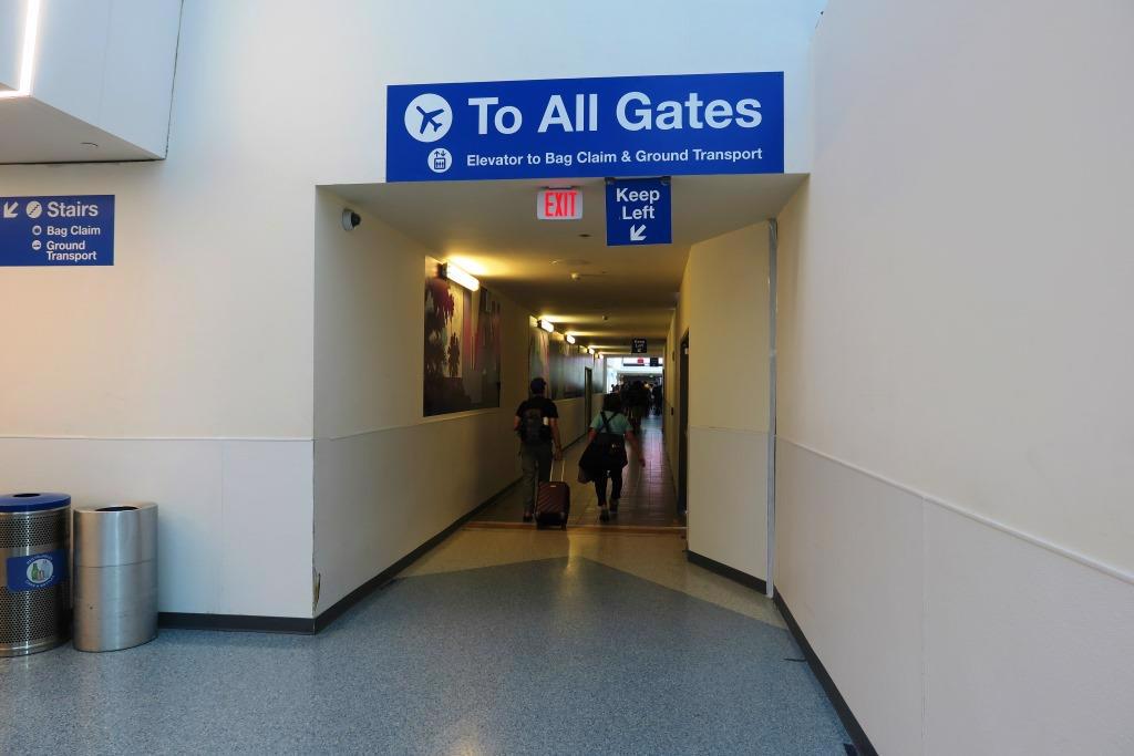 Image of the entrance of a long dark hallway that leads to all gates in terminal 1. There is a small blue sign hanging near the center of that hallway asking pedestrians to keep to the left. Above it is a much bigger sign in the same shade of blue with large white letters informing guests that hallways leads to all gates. On the left of the picture is another small blue sign on the wall telling guests that there are stairs leading to bag claim and ground transport. The stairs are not visible in image. The walls are ivory and white and the floor tiles are a light shade of blue. 