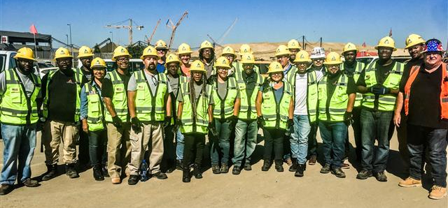 Another group picture consisting of working in yellow reflective vest and yellow hard hats. one worker has an orange vest and an american decorated hardhat. The background has several yellow cranes in the distance where you can also see a large mount of dirt just beyond the machines.