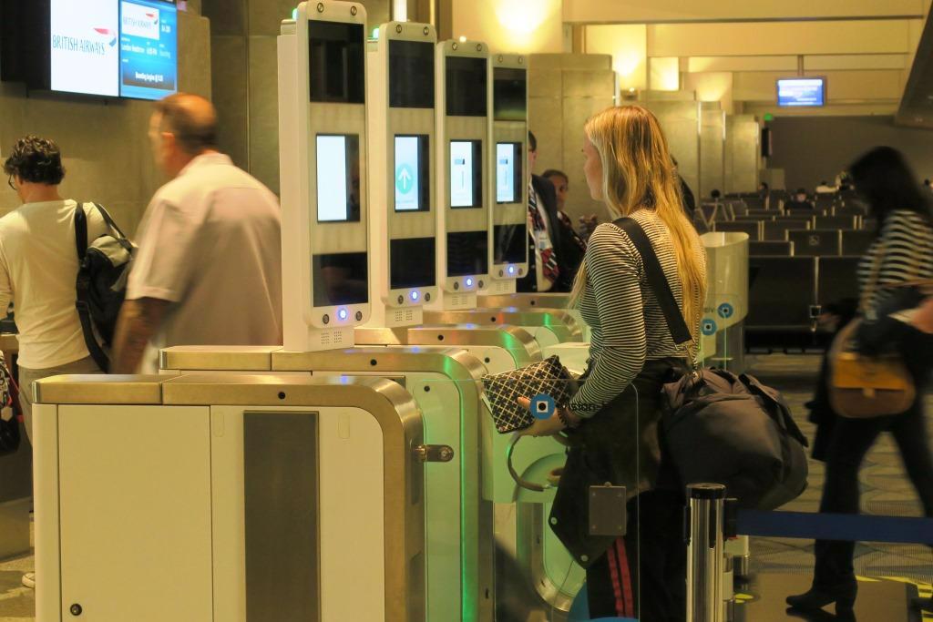  A line of 4 biometric machines with two people going through the completed scanning process, one women approaching machine, and a young lady standing very still as the macine scans her facial features.
