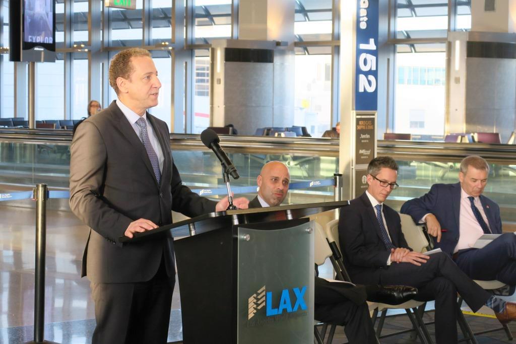 Los Angeles World Airports Chief Innovation and Commercial Strategy Officer Justin Erbacci is seen standing behind a podium with the LAX logo, he is facing an unseen crowd. To his left are three men seated, it is unclear if they have or will be given speeches as well, but they all seem to be listening intently. 
